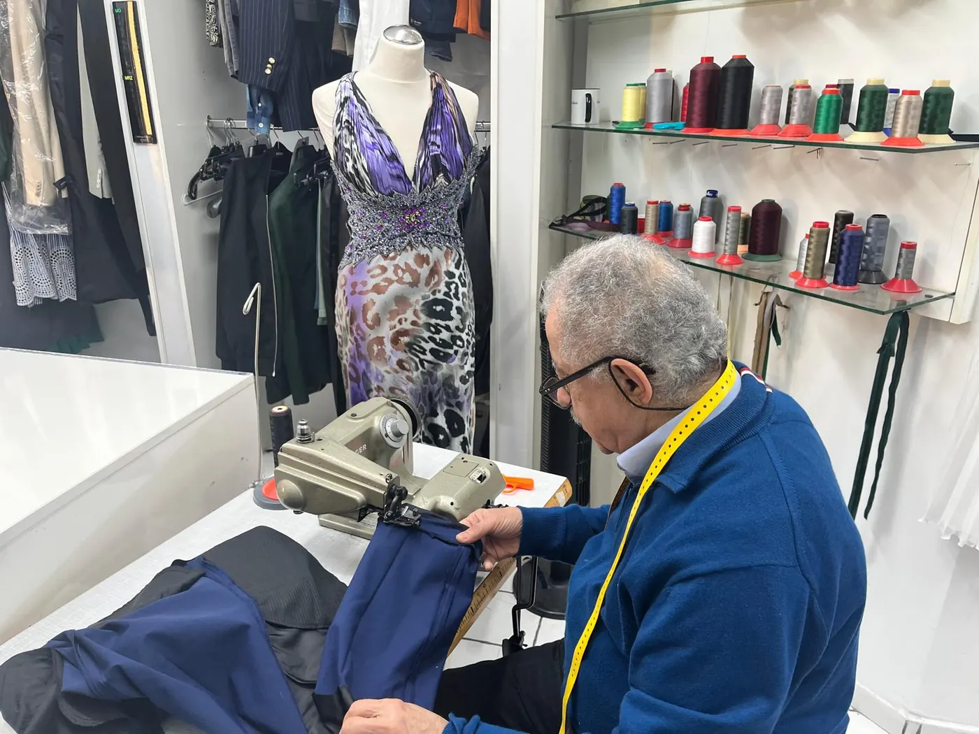 Toni an der JUKI-Industrienähmaschine in der Werkstatt der Änderungsschneiderei Toni in Köln, während er einen Hosenbund anpasst. Im Hintergrund ein Festkleid auf der Schneiderbüste und das Garnrollen-Sortiment.