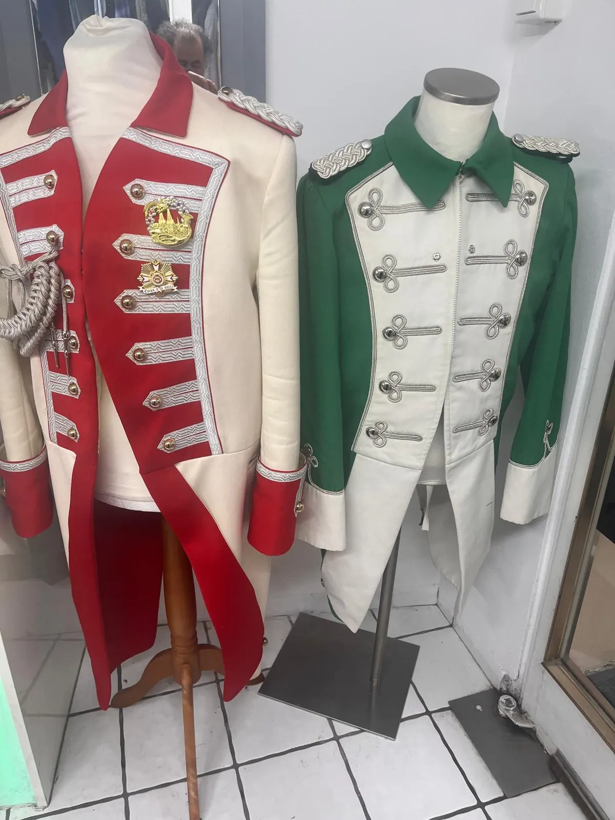 Zwei verzierte Karnevalsuniformen auf Schneiderbüsten in der Werkstatt: rot-creme mit goldener Wappen-Stickerei und grün-creme mit silbernen Knopfreihen.